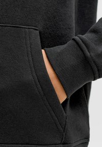 Sudadera negra con capucha, que presenta una tela texturizada, bolsillo canguro, puños de canalé y detalles de costura visibles en los lados y en el dobladillo.