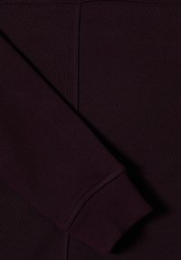 Manica di felpa bordeaux con polsino a costine, caratterizzata da un tessuto testurizzato e cuciture pulite lungo le giunzioni.