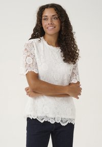 White lace blouse with short sleeves, scalloped hem, and floral patterns. Made of lightweight material, paired with dark denim jeans.