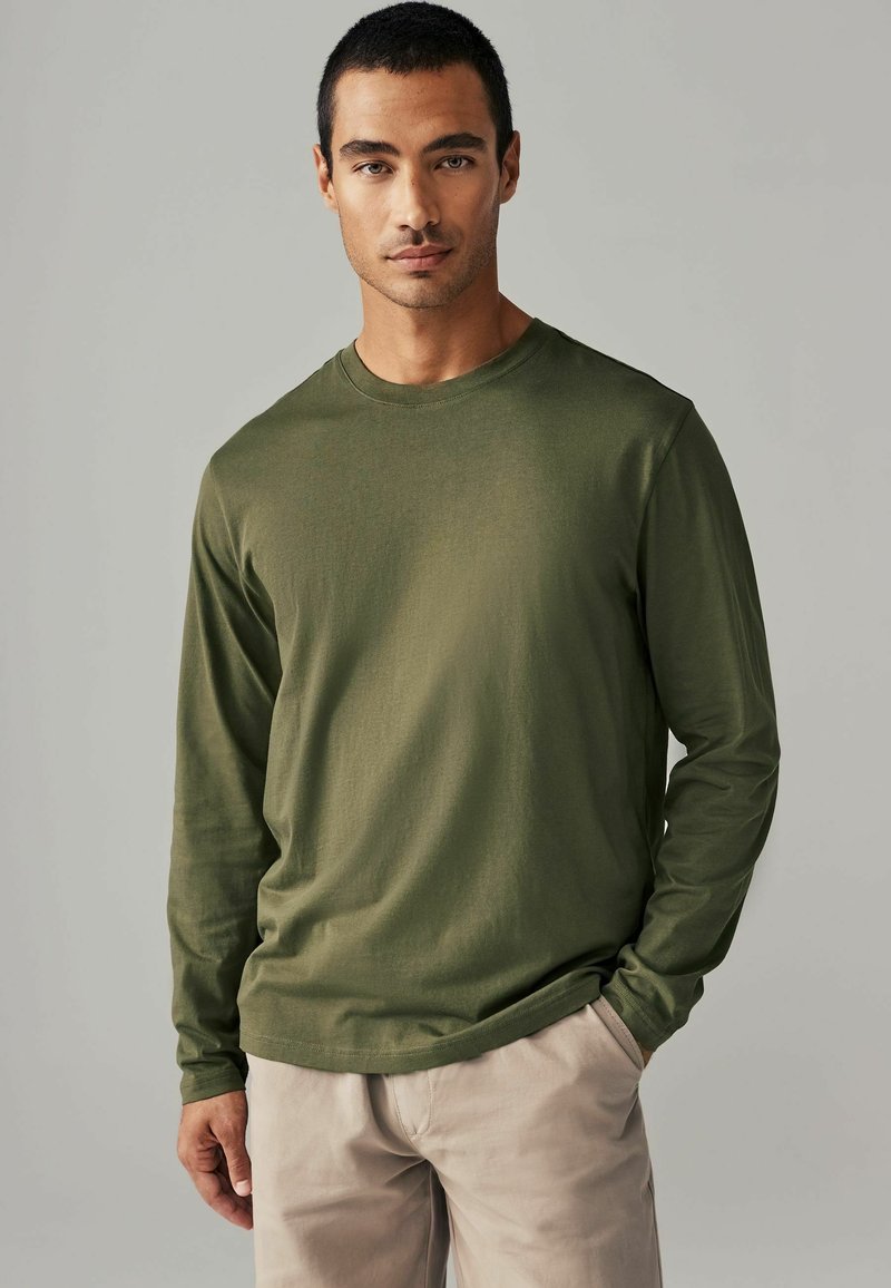 Langärmliges olivgrünes Hemd aus weichem Stoff mit Rundhalsausschnitt, kombiniert mit hellbeigen Hosen. Einfaches Design mit minimalen Akzenten.