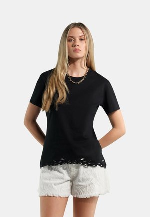 Donna con lunghi capelli biondi che indossa un top nero a maniche corte con dettaglio a taglio sull'orlo e pantaloncini bianchi testurizzati, in piedi con le mani dietro la schiena.