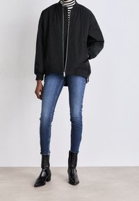 Persoon in een zwarte bomberjack, zwart-wit gestreepte coltrui, blauwe skinny jeans en zwarte enkellaarsjes met hakken, staande op een beige vloer.