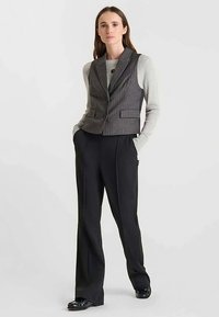 Gilet gris à fines rayures avec une coupe ajustée, porté sur un pull gris clair, associé à un pantalon noir à jambes larges et des chaussures noires brillantes.