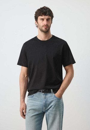 Homme aux cheveux foncés portant un t-shirt noir à manches courtes et un jean bleu clair, debout avec une main dans la poche devant un fond uni.