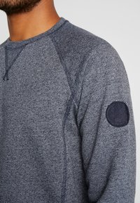 Sudadera de textura gris oscuro con mangas raglán, costuras en contraste y un parche de tela circular en el hombro izquierdo.