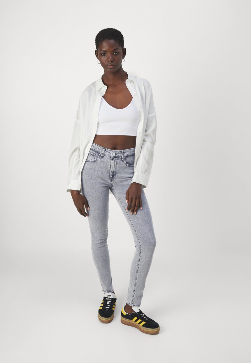 Weißes Hemd über einem weißen Cropped Top, hellgraue Skinny Jeans und schwarze Sneakers mit gelben Streifen, vor einem schlichten Hintergrund.