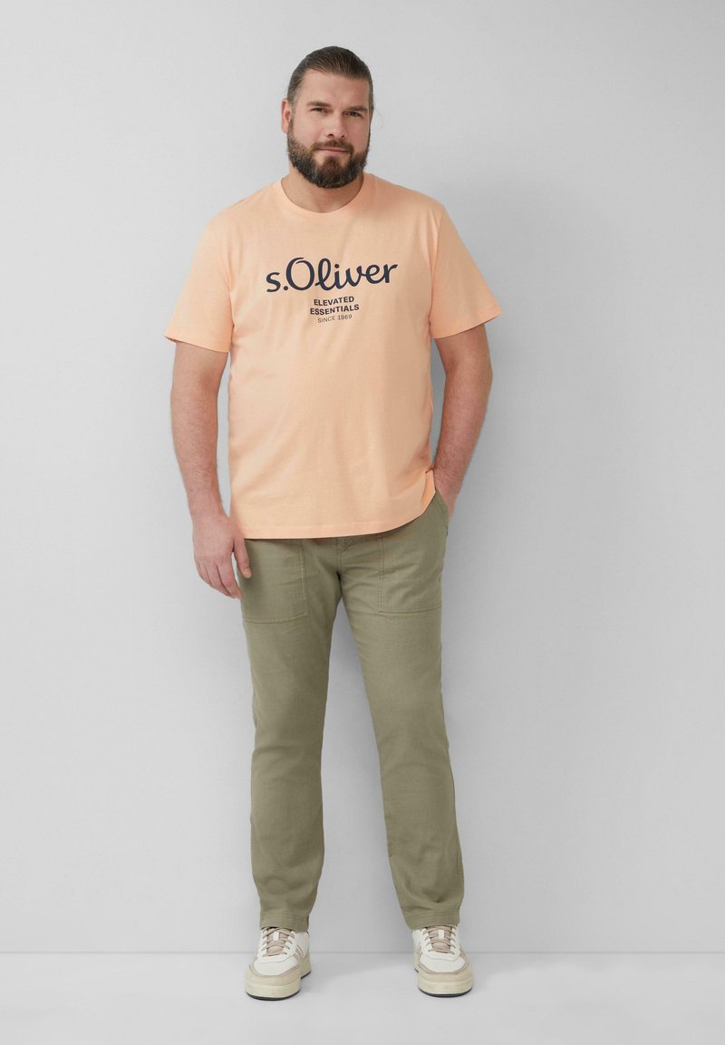 Perzikkleurig katoenen T-shirt met zwarte tekst en logo, gecombineerd met olijfgroene broek en witte sneakers. Eenvoudig, casual ontwerp met een comfortabele pasvorm.