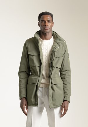 Chaqueta aislante de color verde oliva con cierre de cremallera frontal, cuello alto y cuatro bolsillos frontales. Presenta un suéter de punto texturizado en color beige debajo.