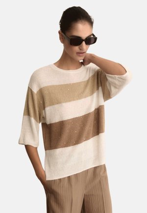 Donna con occhiali da sole neri, maglione largo a righe beige e bianco con brillantini e pantaloni plissé marrone chiaro, in posa con una mano sul collo.