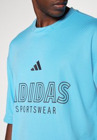 Mėlynos spalvos Adidas sportinė marškinė su tekstūruotu audiniu, priekyje su juodu logo ir užrašu. Atsipalaidavęs siluetas, apvalus kaklas.