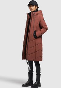 Kvinde iført en rustfarvet lang polstret frakke med hætte, sort kasket, sorte leggings med tekst og sorte ankelstøvler, stående på siden.