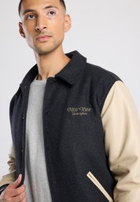 Dark wool-blend varsity jacket with cream leather sleeves, featuring "NEW YORK" embroidered in gold on the chest and snap button closure.