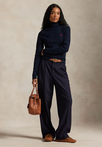 Γυναίκα που φοράει μπλε ζιβάγκο, φαρδιά παντελόνια, καφέ ζώνη και παπούτσια, κρατώντας καφέ δερμάτινη τσάντα σε απλό φόντο.