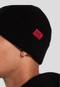 Primo piano di una persona che indossa un berretto a coste nero con un'etichetta rossa "WRONG FRIENDS" e un piccolo orecchino a cerchio nell'orecchio sinistro.