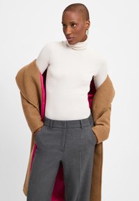 Beige coltrui, bruine wollen jas met een roze voering, grijze op maat gemaakte broek met zijzakken, model staat in een ontspannen pose.