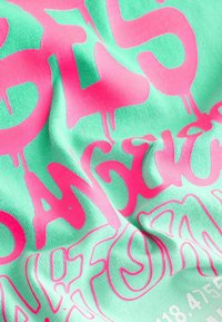 Mátově zelená látka s výrazným růžovým textem se slovem "NEBEZPEČNÉ" a abstraktními vzory. Materiál vypadá měkce s mírně texturovaným povrchem.