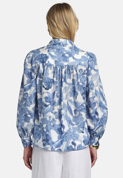 Woman with blonde hair wearing a blue and white floral long-sleeve blouse and white pants, viewed from the back.