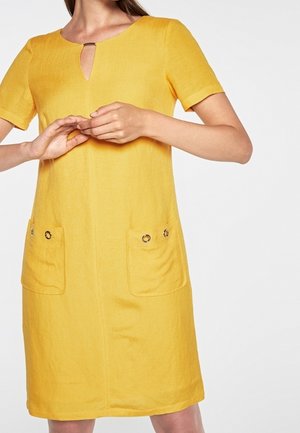 Mujer con un vestido amarillo de manga corta, con un pequeño escote en forma de ojo de cerradura y dos bolsillos delanteros con detalles de botones.