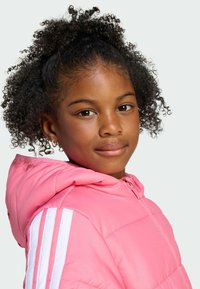Young girl with curly hair smiles while wearing a pink puffer jacket with white stripes, showcasing her friendly expression and playful style.