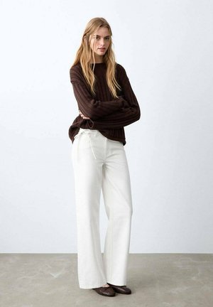 Pull en tricot côtelé marron, pantalon blanc à jambes larges, ballerines marron. Le mannequin est debout, les bras croisés, devant un fond clair.