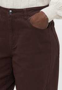 Hnědé bavlněné kalhoty se rovnýma nohama, s bočními kapsami a kovovým zapínáním v pase, viditelné švy.