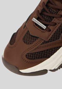 Sneaker marrone in materiale sintetico liscio, con pannelli in rete, lacci testurizzati e una suola intermedia bianca con una suola esterna in gomma resistente. Dettaglio di marchio in argento.