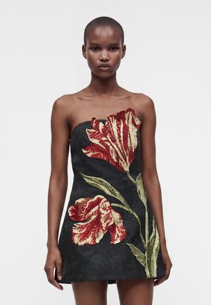 Mannequin portant une mini-robe noire bustier avec une grande broderie florale rouge et crème et des tiges vertes à l'avant, debout devant un fond uni.