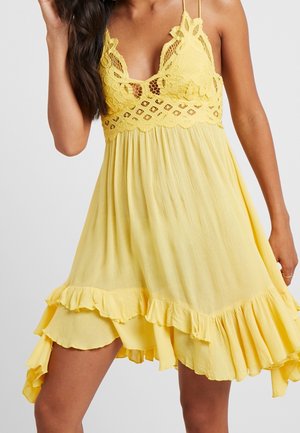 Femme portant une robe jaune sans manches avec un corsage en dentelle, des bretelles ajustables et un ourlet asymétrique à volants.