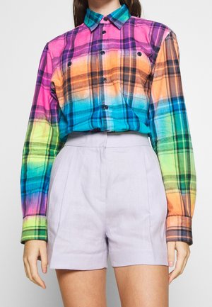 Vrouw draagt een kleurrijk geruit overhemd met een regenboogverloop, ingestopt in hoge lichtlavendelkleurige shorts tegen een effen achtergrond.