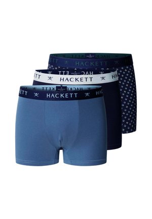 Jonge man staat op blote voeten tegen een effen achtergrond en draagt blauwe boxershorts met een merkbare tailleband.
