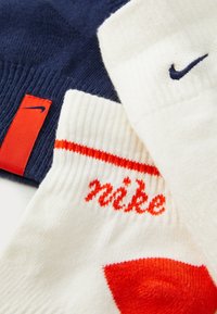 Tres calcetines: uno azul marino con etiqueta roja, uno blanco con una franja naranja y texto "nike", y un calcetín blanco con un pequeño logo azul.