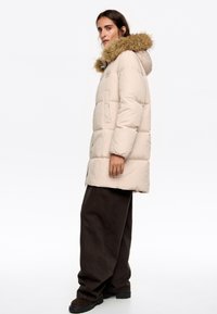 Chaqueta tipo plumas en beige claro con capucha de piel sintética. Presenta un diseño acolchado, bolsillos frontales y un corte recto. Combinada con pantalones marrón oscuro.