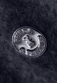 Insignia circular de plata con la silueta de un perro, con el texto grabado "FRIEDA & FREDDIES" sobre un fondo de tela oscura texturizada.