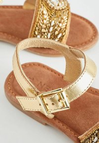 Goldene metallische Sandalen mit perlenverzierten Akzenten, weichem braunem Wildlederfußbett, verstellbarem Knöchelriemen mit metallischer Schnalle und flacher Sohle.