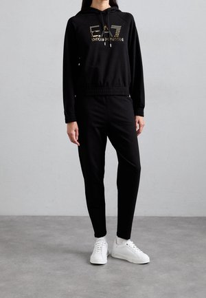 Personne portant un sweat à capuche noir avec le logo doré EA7 Emporio Armani, pantalon noir et baskets blanches, debout sur un sol gris contre un mur clair.