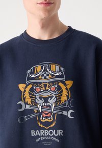Navy sweatshirt featuring a graphic of a tiger wearing goggles, holding wrenches. The design includes bold colors and detailed facial features.