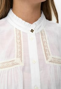 Primo piano di una camicia bianca con bordo di pizzo, colletto arricciato e un piccolo bottone a forma di fiore nero e oro al collo.
