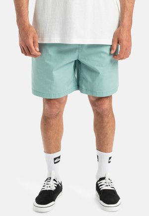 Shorts bleu clair avec des coutures visibles, fabriqués en tissu coton doux. Portés avec des baskets noires et des chaussettes blanches arborant un logo.