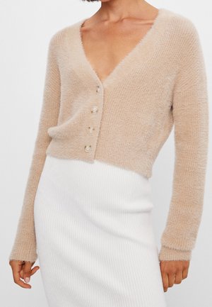 Béžový chlupatý kardigan se čtyřmi knoflíky nošený přes bílou žebrovanou sukni, zobrazený od ramen po boky, s pažemi uvolněně podél těla.