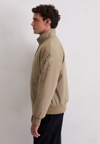 Jasnobrązowa kurtka z wysokim kołnierzem, zamkiem błyskawicznym, elastycznymi mankietami i talią. Posiada boczną kieszeń oraz małą naszywkę z logo na rękawie.