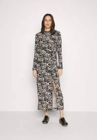 Vestido de manga longa com padrão preto e branco, com uma fenda lateral, feito de tecido suave. Modelo usa tamancos pretos com salto em madeira.