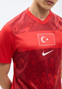 Άνδρας που φοράει κόκκινη αθλητική φανέλα με την τουρκική σημαία και λευκό λογότυπο της Nike, με μαρμαρυγιακό μοτίβο στο στήθος και στους ώμους.