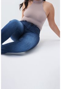 Mujer con jeans ajustados azules de talle alto y un top sin mangas color beige estilo halter, sentada con un brazo apoyado en el suelo blanco.