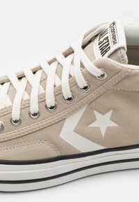 Beige mockasinsneakers med vit stjärnlogotyp, vita snören och vit gummisula med svart rand, märkt All Star på den vadderade tungan.