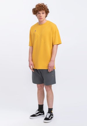 T-shirt gialla oversize con dettagli di cuciture diagonali, abbinata a pantaloni corti grigi e scarpe nere con accenti bianchi.