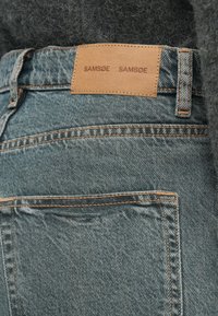 Gaiši zili džinsi no denima ar ādas birku, uz kuras rakstīts "SAMSOE SAMSOE." Iezīmē oranžs diegs un aizmugurējās kabatas ar vienkāršu dizainu.