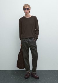 Junger Mann in einem dunkelbraunen Pullover, schwarzen Jeans, braunen Schuhen und Sonnenbrille, der eine dunkle Tasche hält und vor einer schlichten hellgrauen Wand steht.