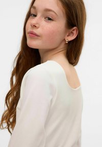 Top bianco a maniche lunghe con design a scollo profondo sul retro. La modella ha capelli castani mossi e indossa piccoli orecchini a bottone in oro. Tessuto dalla texture liscia.