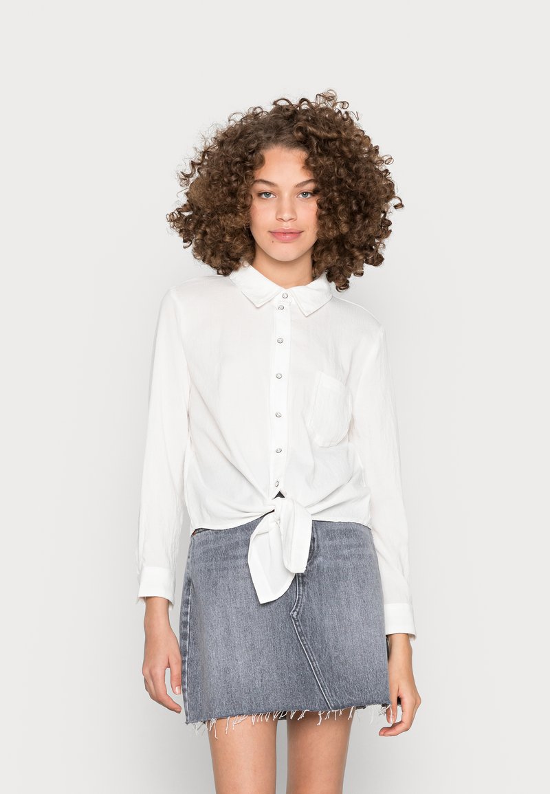 Witte, knoopsluiting blouse met een gestrikte voorkant en een enkele borstzak, gecombineerd met een grijze denimrok met rafelige randen.