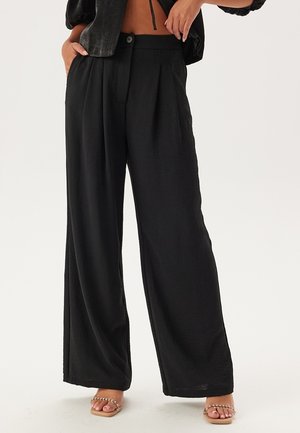 Pantalons larges noirs en tissu léger, avec des plis, une fermeture à bouton et une texture lisse. Associés à des sandales beiges.